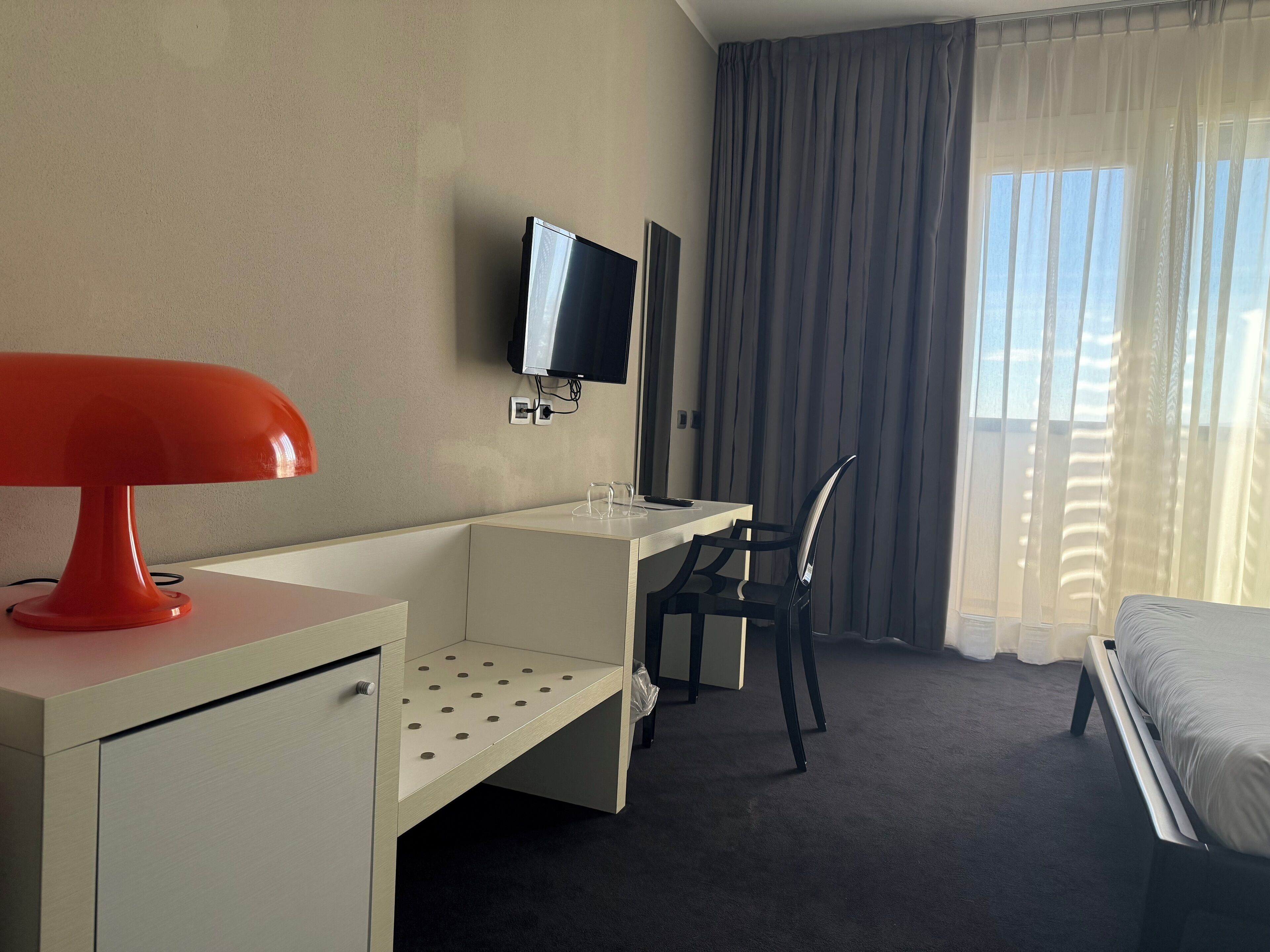 Une chambre d’hôtel équipée d’un bureau, d’une chaise, d’un lit, d’une télévision et d’une fenêtre avec des rideaux.