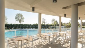 Un espace piscine extérieur abrité, doté de chaises longues et de tables, offrant une vue dégagée sur la piscine et s’intégrant dans un décor paysager.