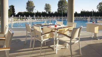 An outdoor pool area with lounge chairs and tables, surrounded by trees and a clear sky.