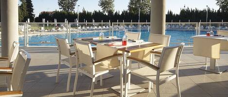 Ett utomhusområde med en pool, solstolar och bord, omgivet av träd och en klar himmel.