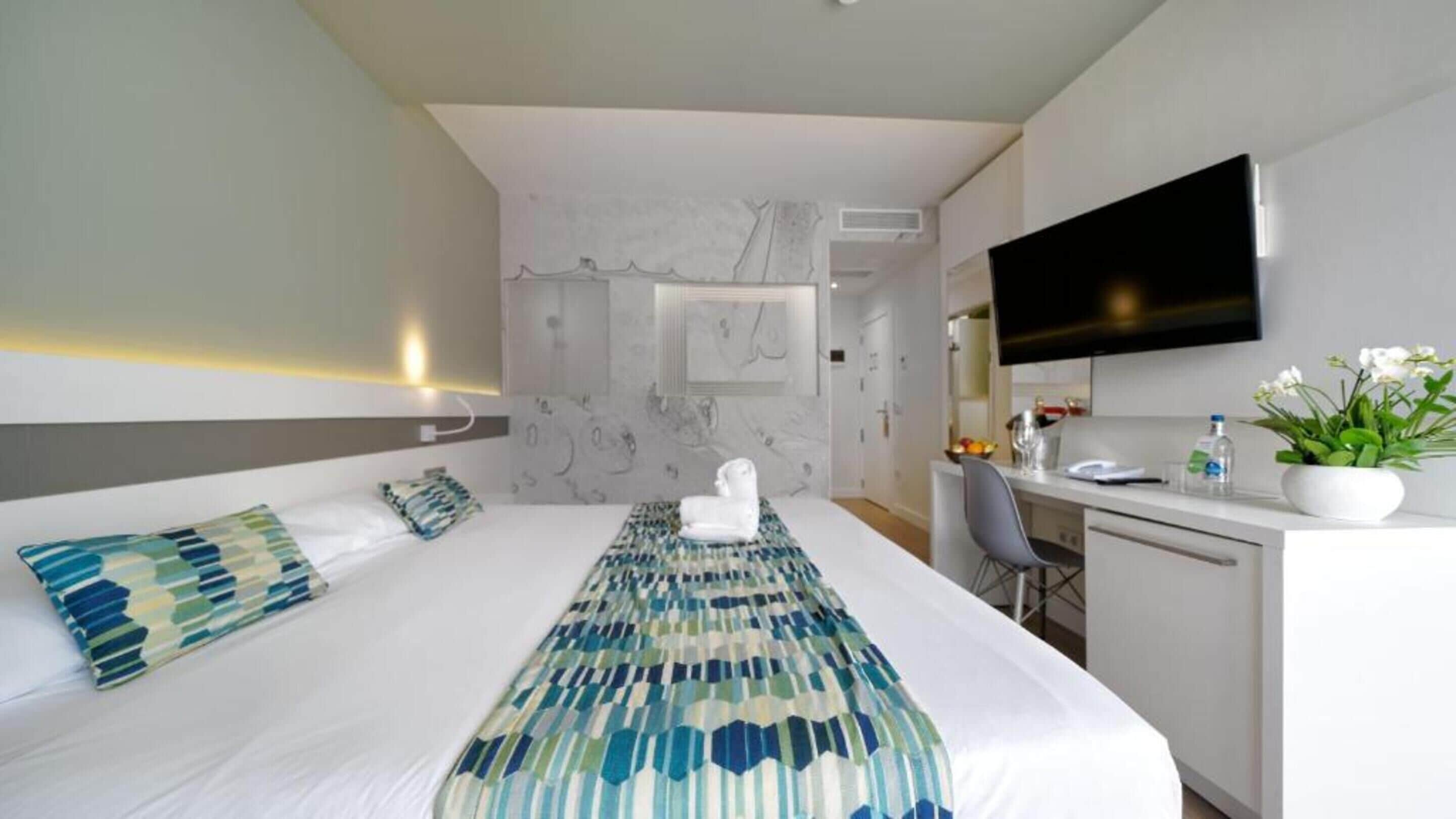 Quarto de hotel com uma cama grande, uma escrivaninha com televisão de tela plana, uma cadeira e um vaso de planta.