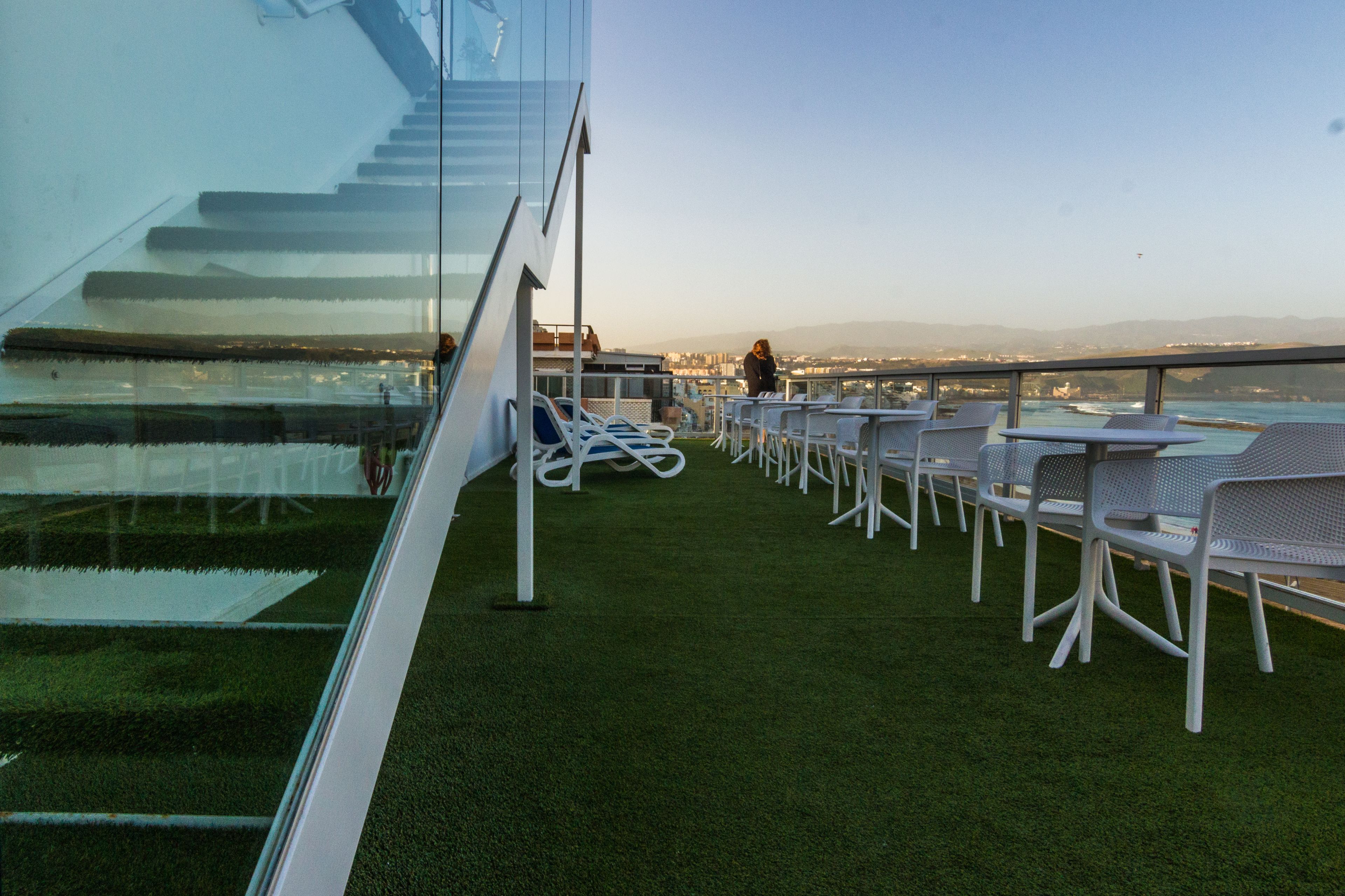 Eine Dachterrasse mit künstlichem Gras, weißen Metallstühlen und einer Glaswendeltreppe.