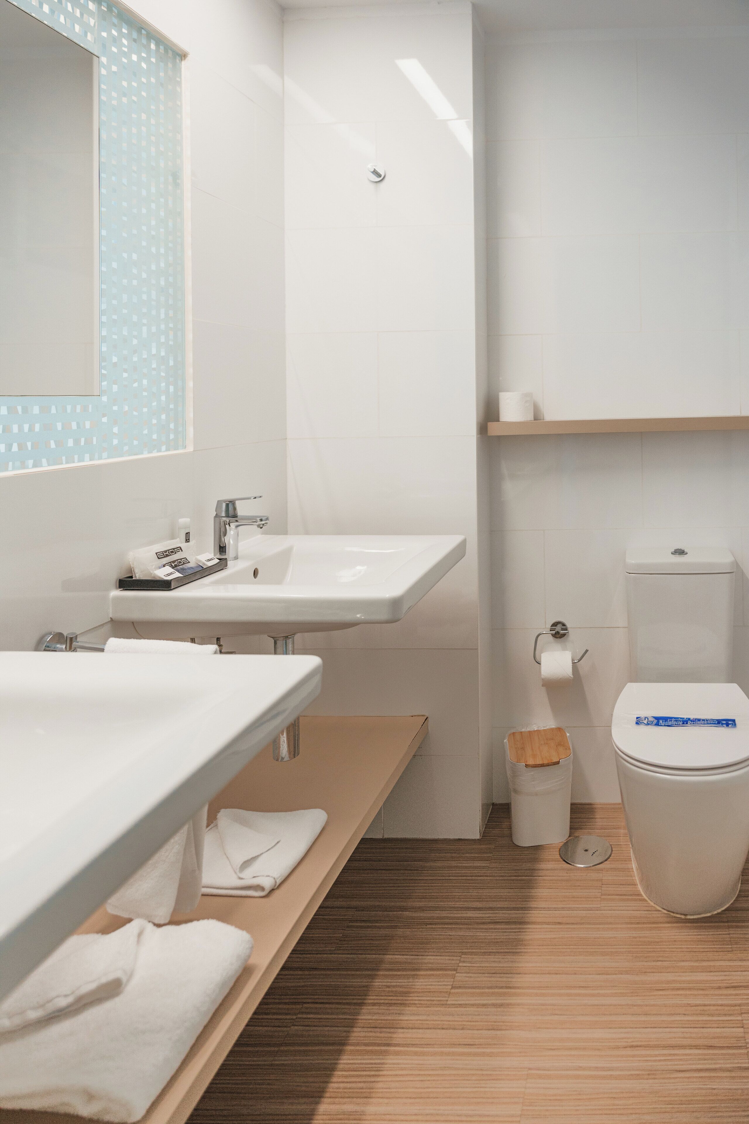 A modern bathroom with a wooden vanity, a white sink, a toilet, and a trash bin.