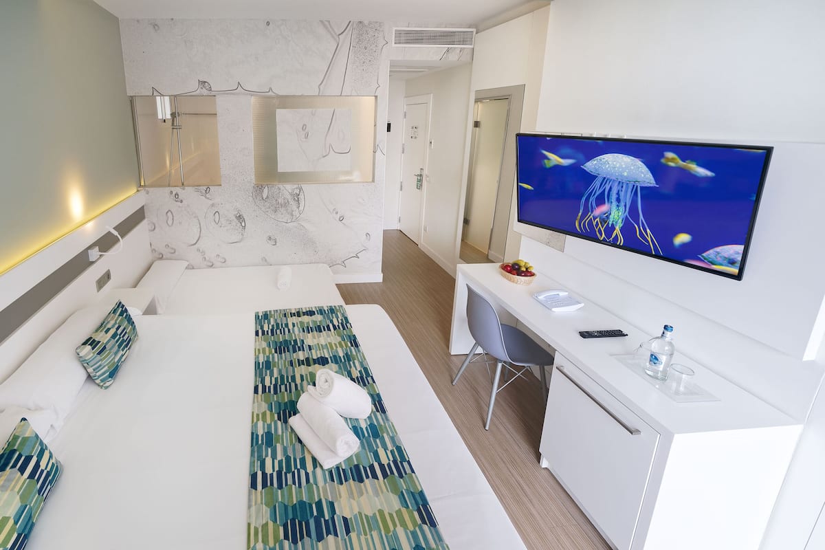 A modern hotel room with a large bed, a desk with a computer, and a TV displaying a jellyfish.
