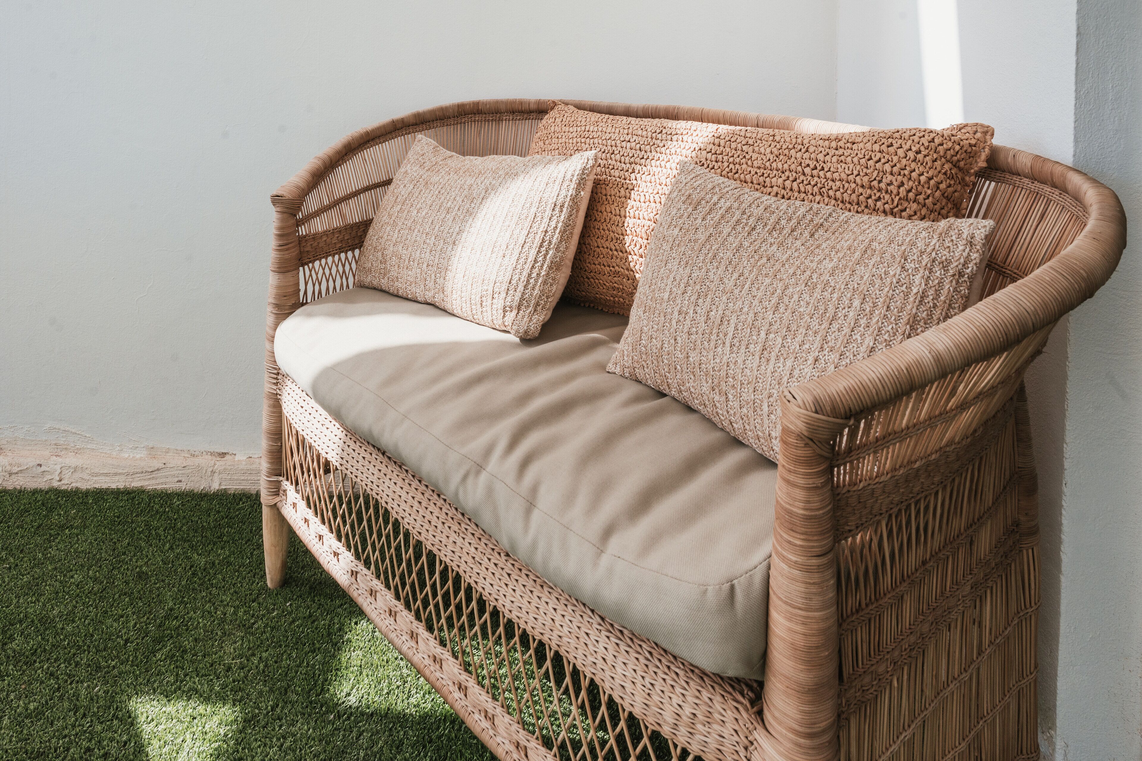 A wicker sofa with beige cushions against a white wall, positioned on a patch of artificial grass.