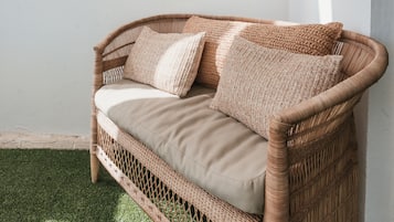A wicker sofa with beige cushions against a white wall, positioned on a patch of artificial grass.