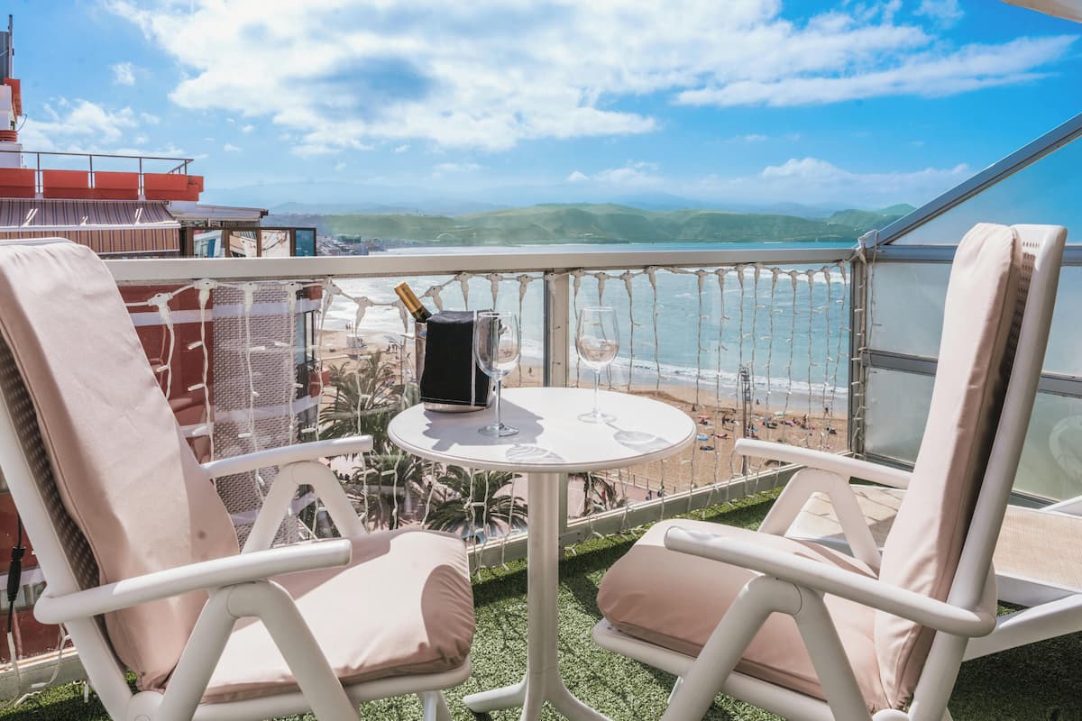 A balcony with two white cushioned chairs, a small round table, and a bottle of champagne. The view includes a beach and distant hills.