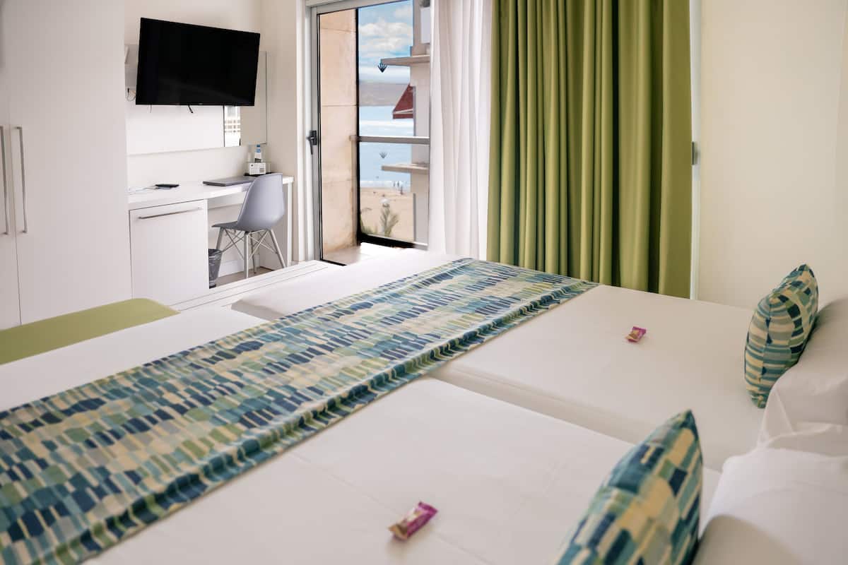 A hotel room with a bed, a desk with a chair, a television, and a view of the beach through a sliding door.