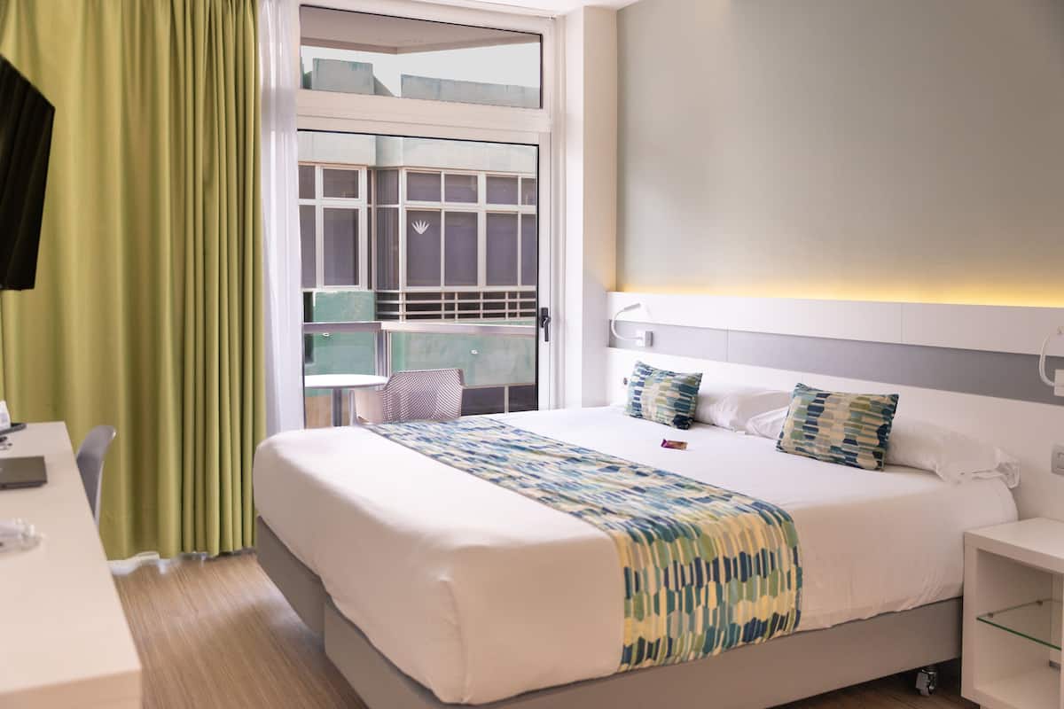 A hotel room with a large bed, a desk, a chair, a window with curtains, and a bedside table.