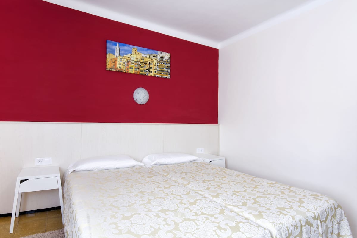 Een slaapkamer met een rode accentmuur, een bed met een geborduurd beddegoed, een nachtkastje en een klok aan de muur.
