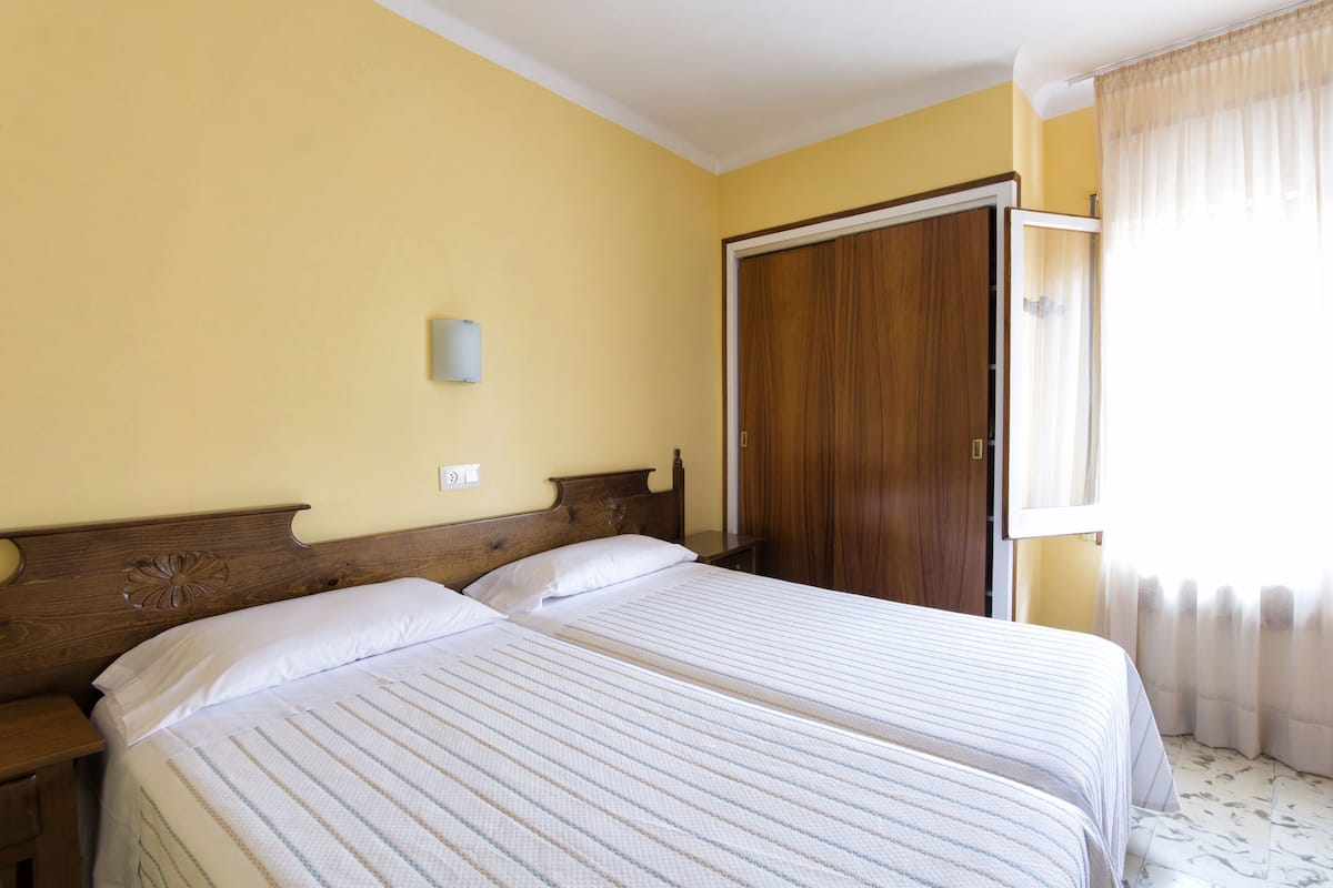 Hotelkamer met twee bedden, houten hoofdeinden, een nachtkastje, een kledingkast en een raam met gordijnen.