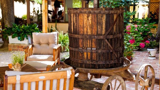 Un espace extérieur rustique aménagé avec un tonneau en bois muni de roues, entouré de plantes en pot et agrémenté d’un fauteuil rembourré.