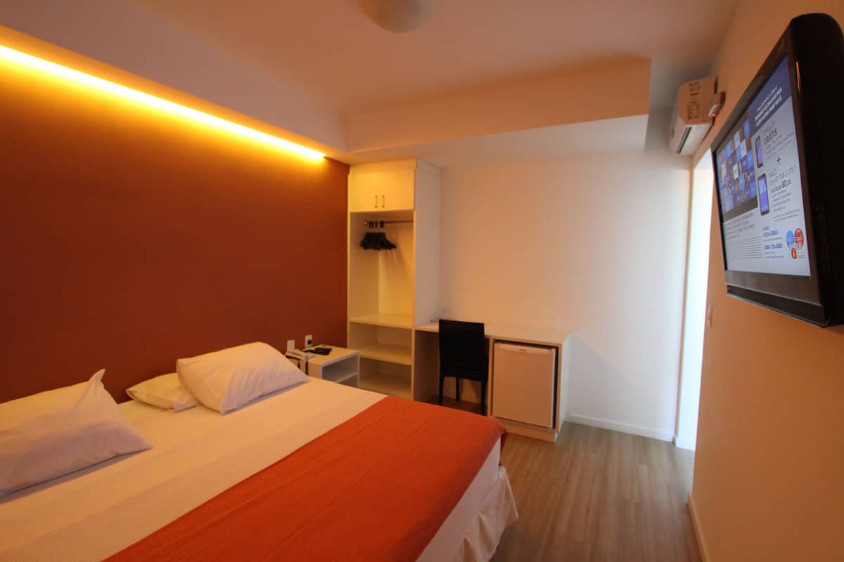 A hotel room with a bed, a desk, a chair, a small fridge, and a wall-mounted TV.