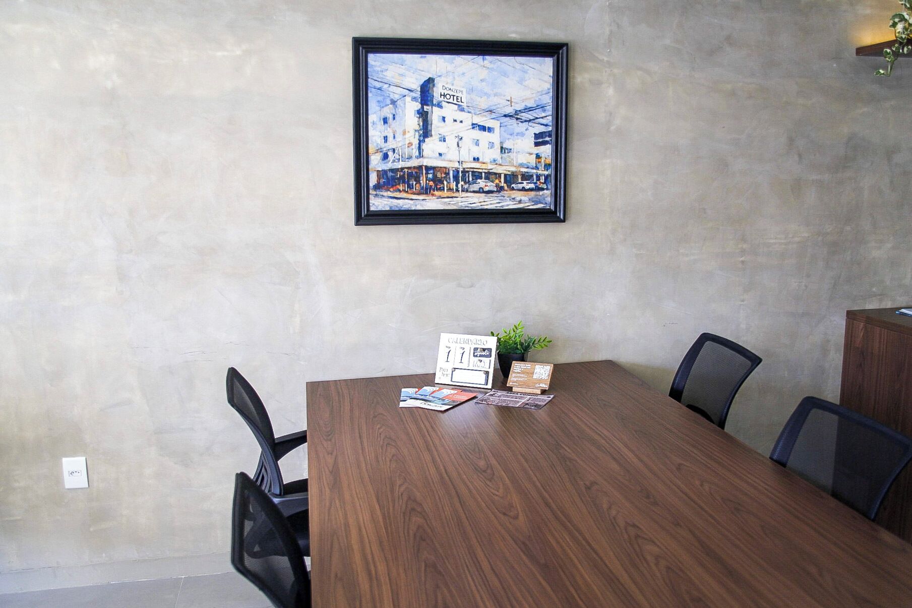 Un salón de conferencias con una mesa de madera, sillas, un cuadro enmarco, un calendario y una pequeña planta en maceta.