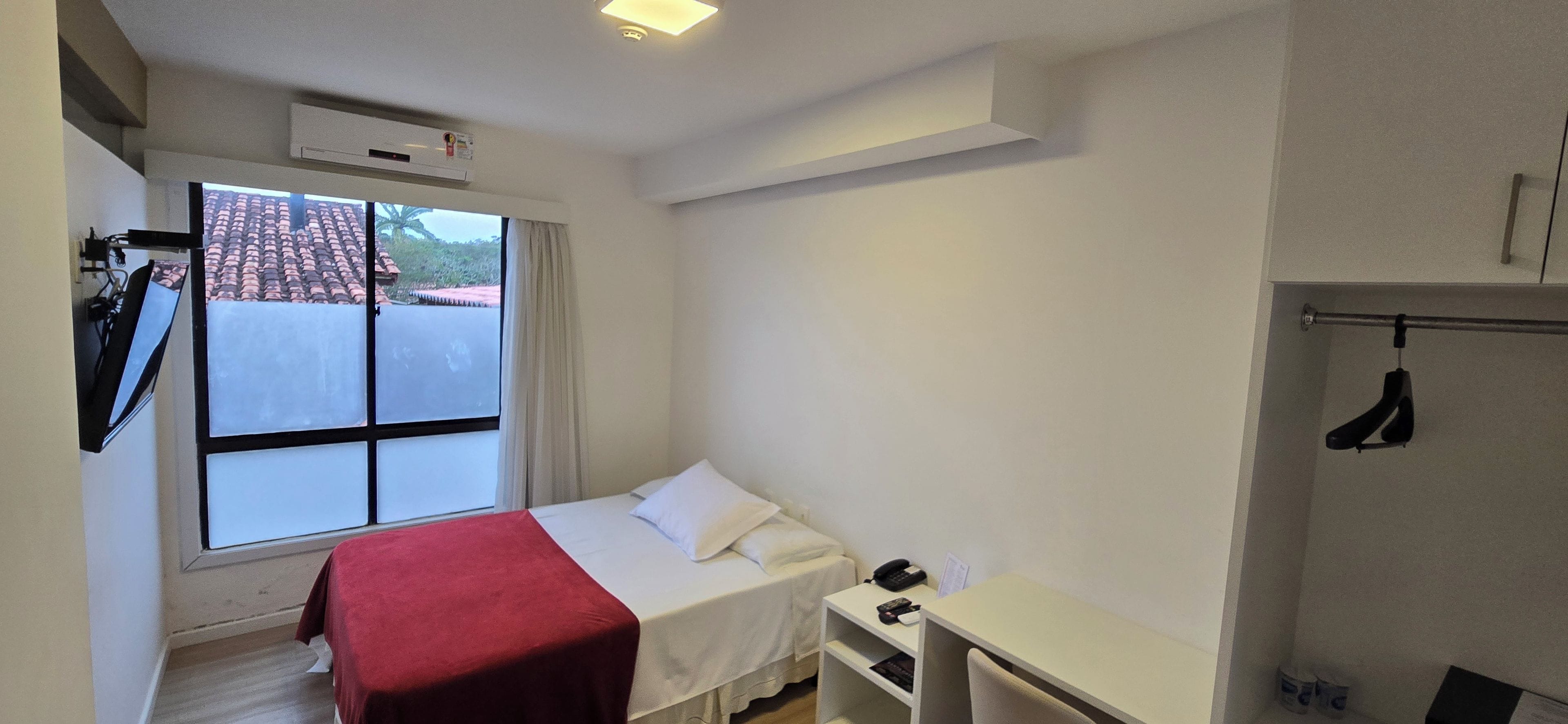 Una habitación pequeña y limpia con cama, escritorio, silla, ventana con vistas a edificios y perchero.