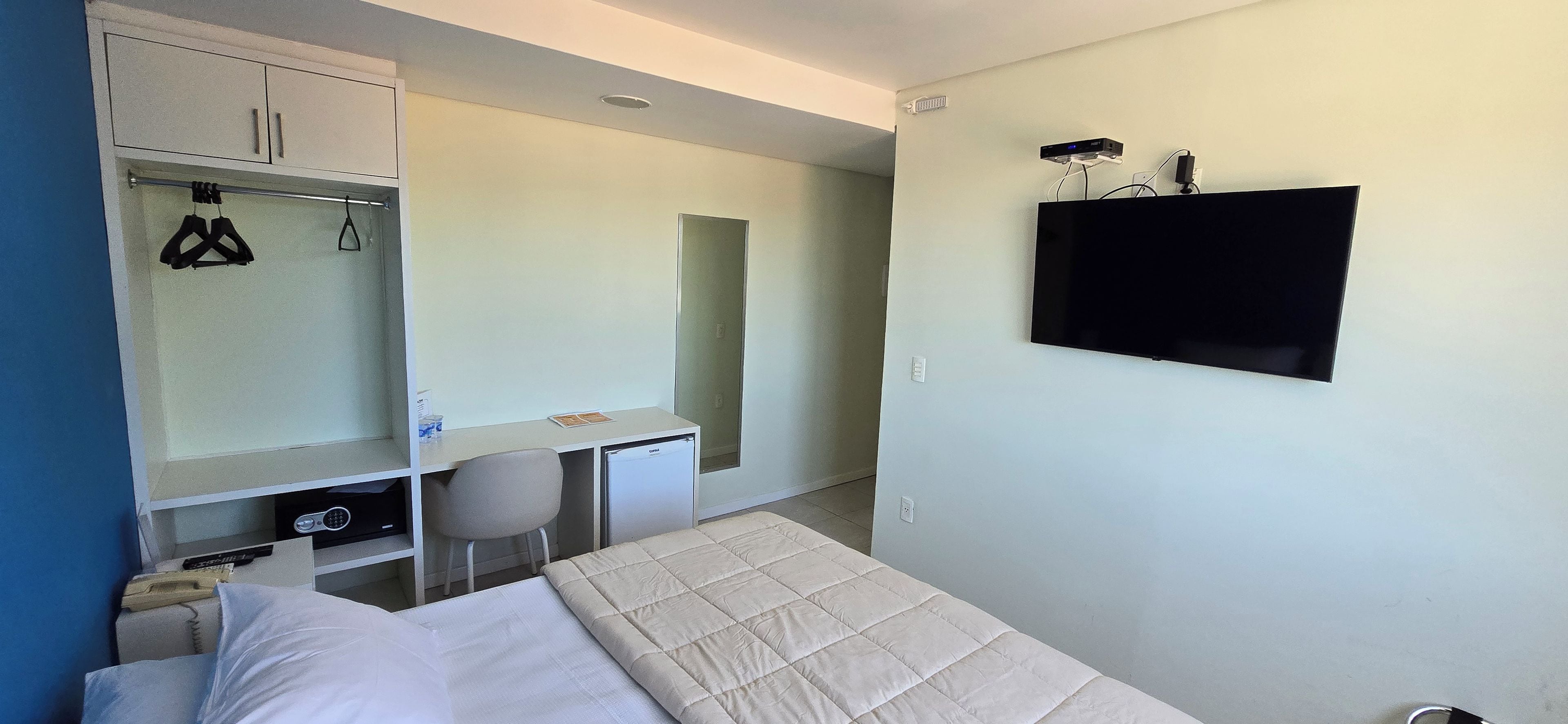 Ein Schlafzimmer mit einem Bett, einem Schreibtisch und einem Stuhl. Ein Kleiderschrank mit Kleidung und ein an der Wand befestigtes Fernsehgerät.