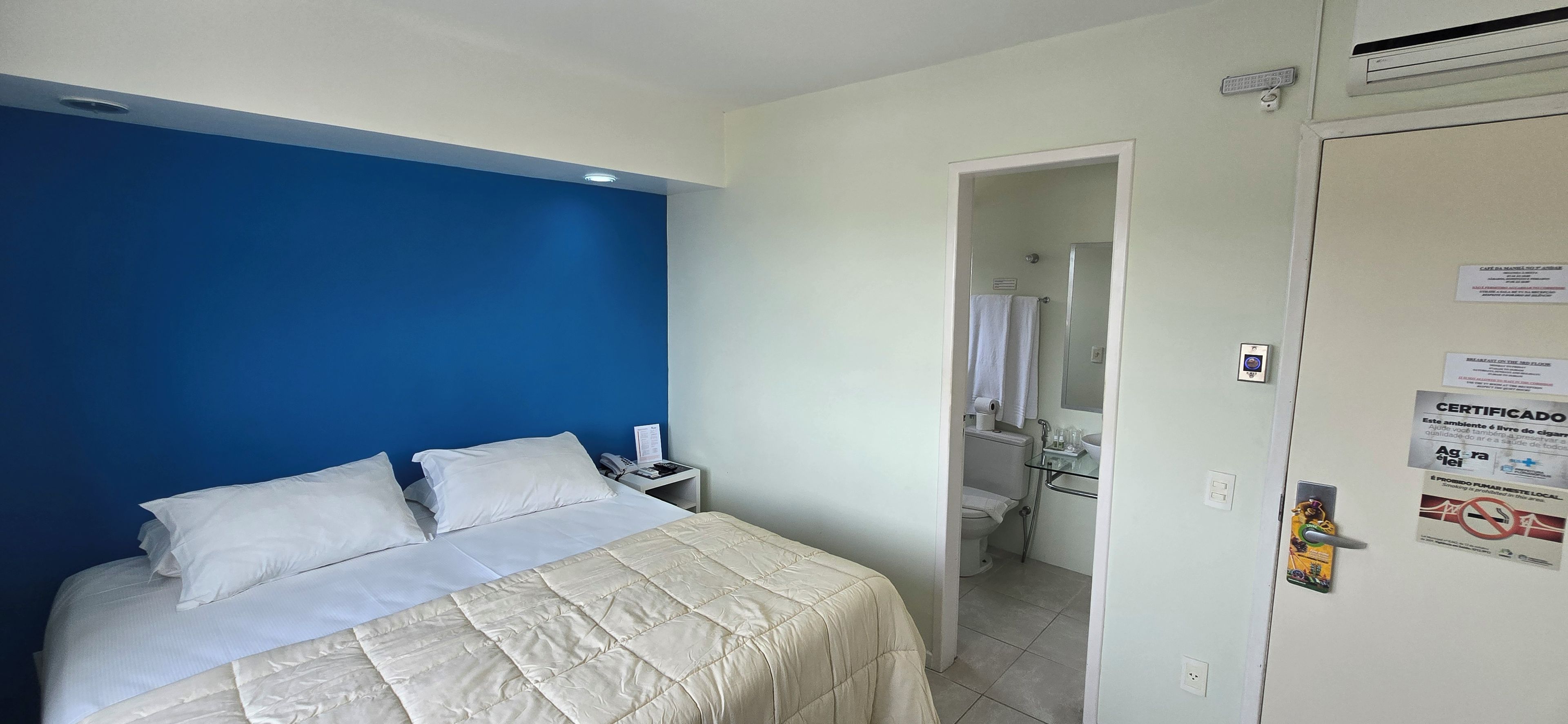 A hotel room with a bed, a nightstand, a bathroom visible through an open door, and a wall with a blue accent.