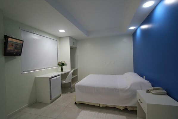 A bedroom with a bed, a desk, a chair, a small cabinet, and a wall-mounted TV.