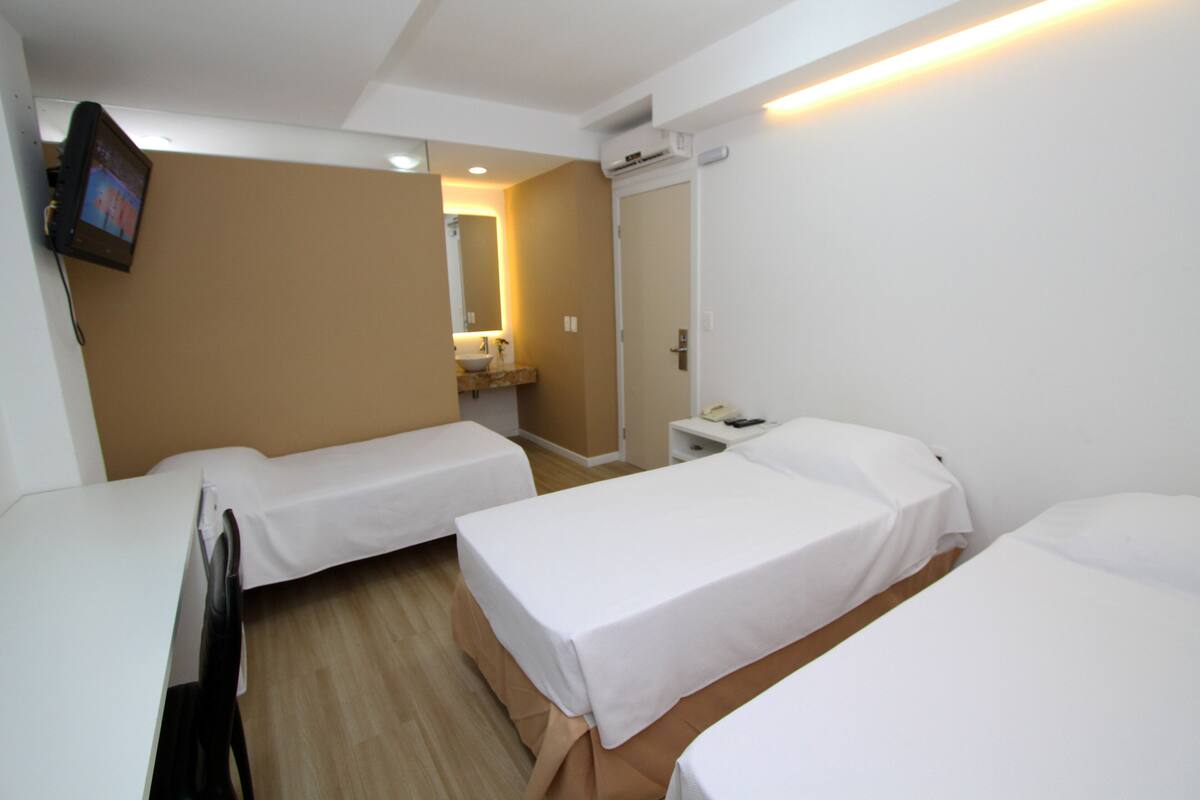 A hotel room with two single beds, a small bedside table, a nightstand, a TV mounted on the wall, and a bathroom with a sink and mirror.