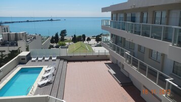 Piscina en la azotea con sillones de descanso, vista al mar y un edificio moderno con grandes ventanales.
