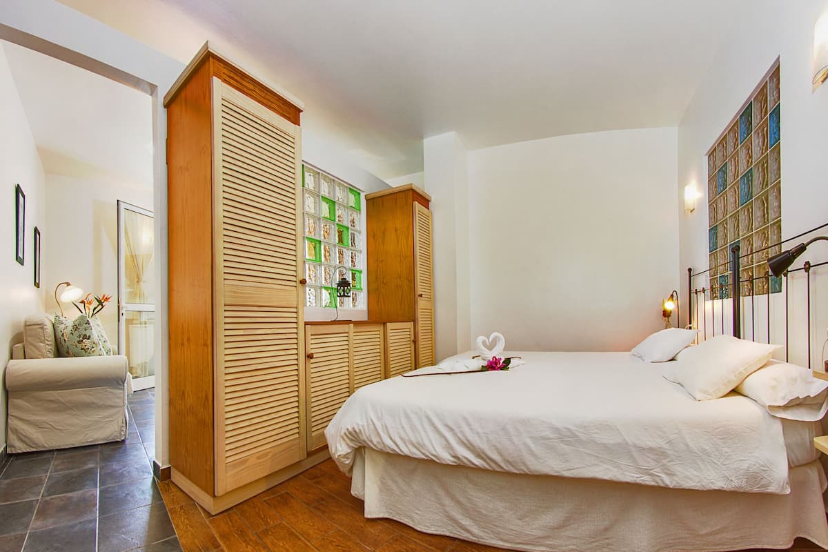 A bedroom with a large bed, wooden wardrobe, and a small seating area with a lamp.