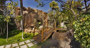 A wooden bridge over a small water feature, surrounded by lush greenery and a large rock formation. - Adjovimar Apartamentos (Los Llanos de Aridane)