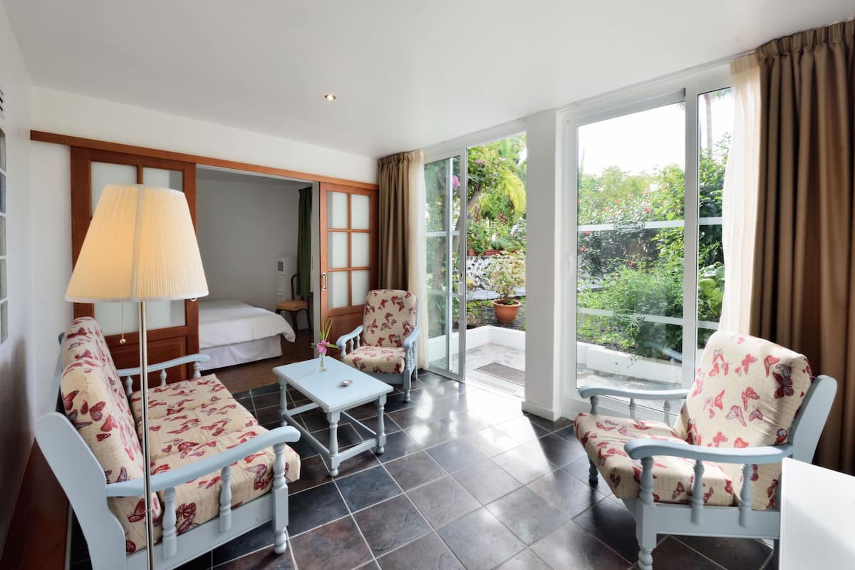 A hotel room with a bed, two chairs, a table, a lamp, and a large window offering a view of the garden.
