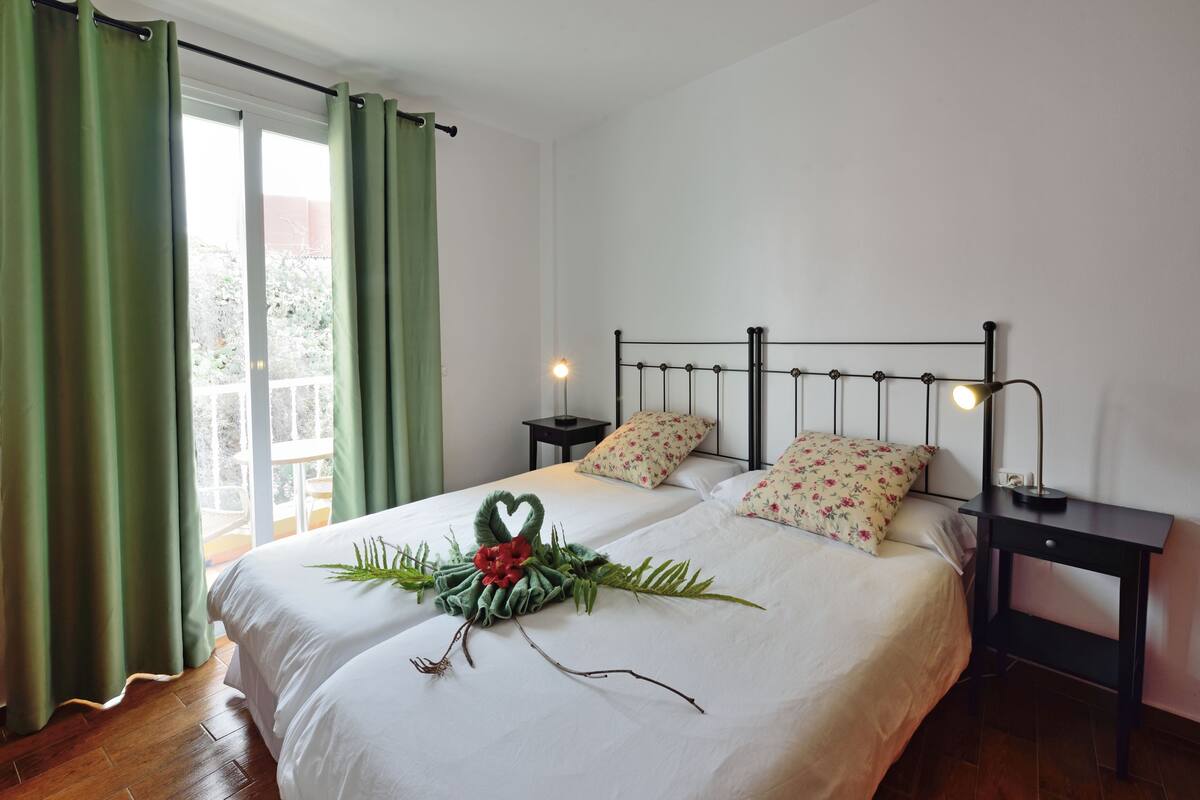 A neatly made bed with a heart-shaped decoration, a nightstand with a lamp, and a view of greenery through the window.