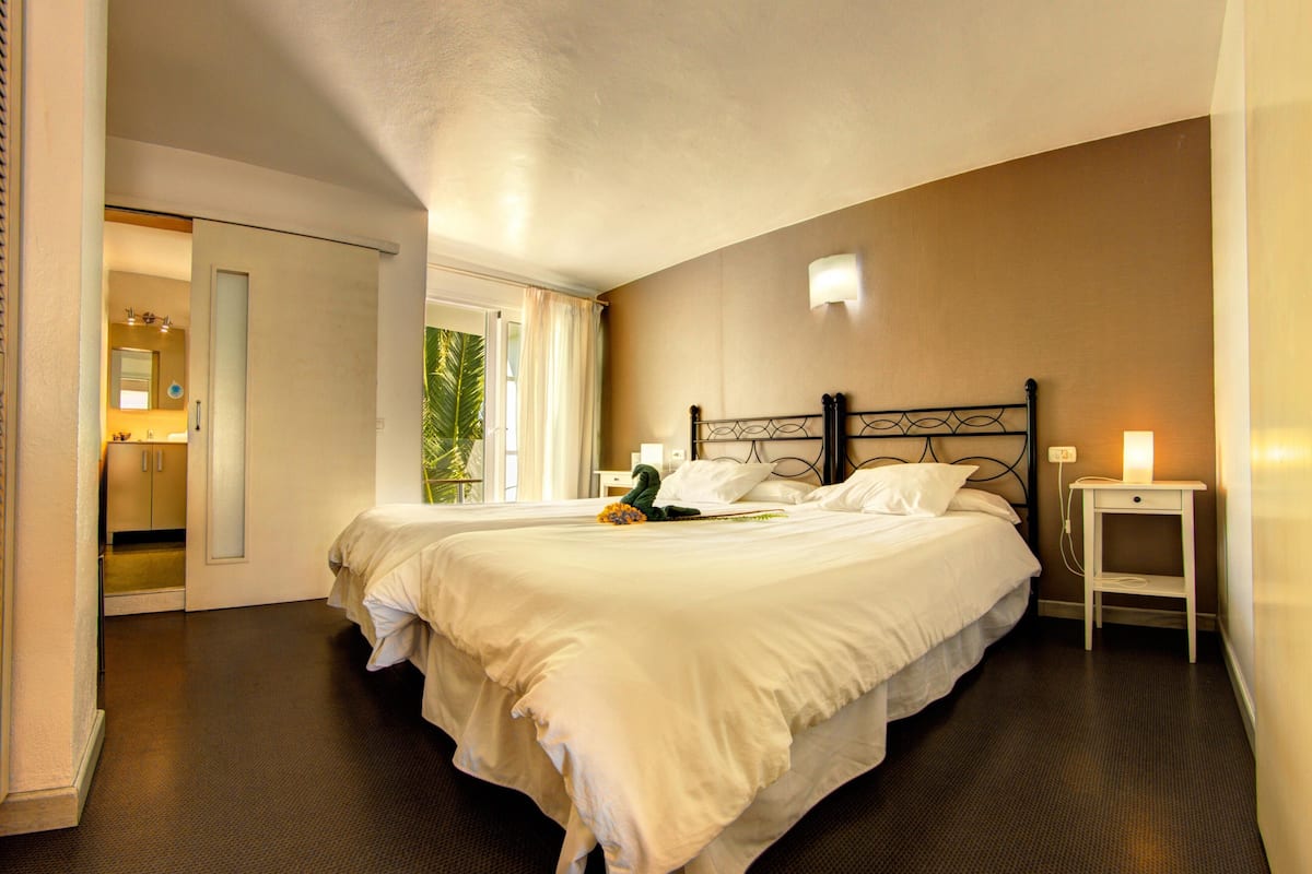 A large bed with white linens, a bedside table with a lamp, and a view of a bathroom with a mirror and sink.