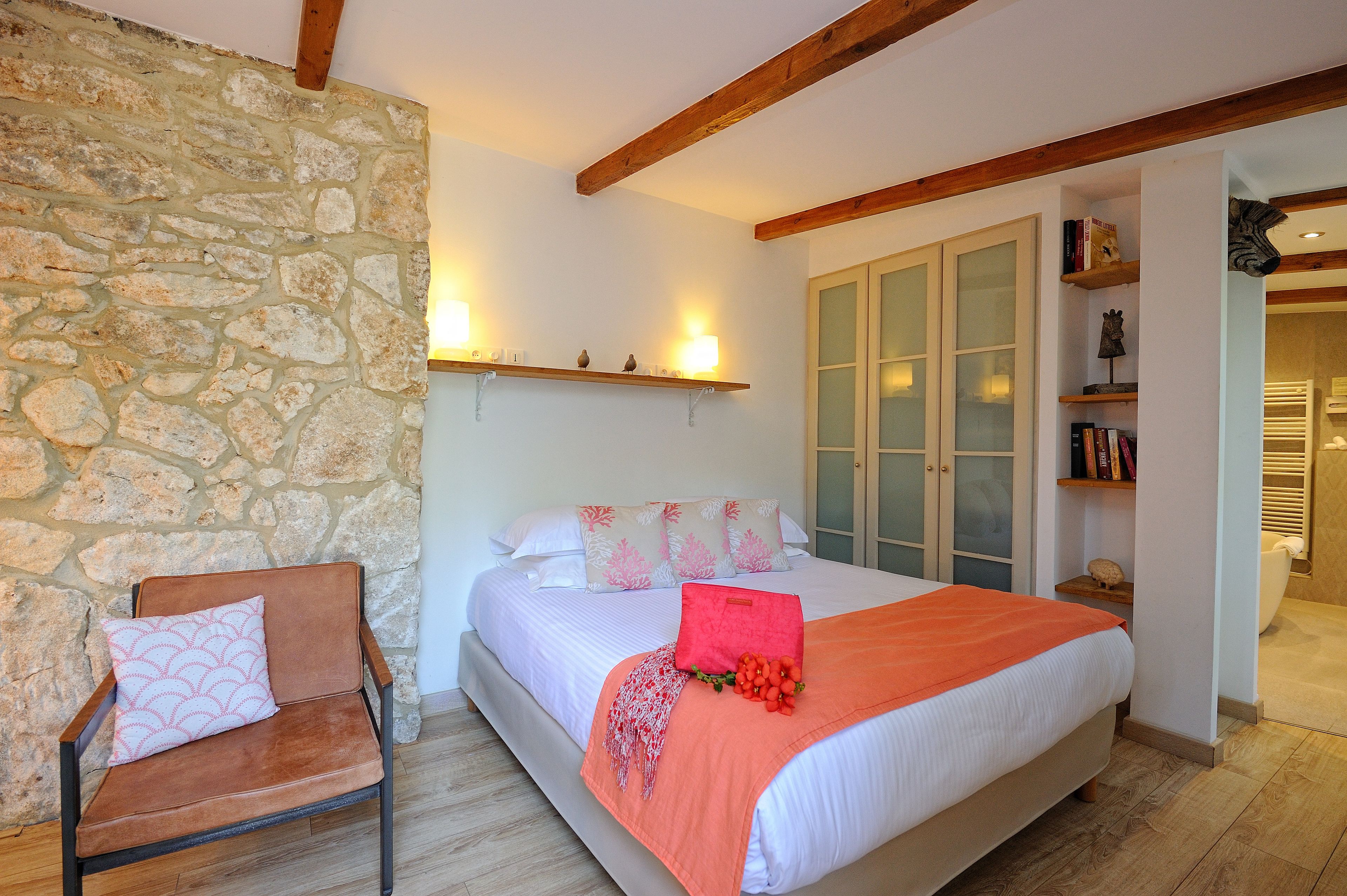 Een slaapkamer met een stenen muur, een bed met wit beddengoed en roze kussens, een houten stoel, en een plank met boeken en decoratieve objecten.