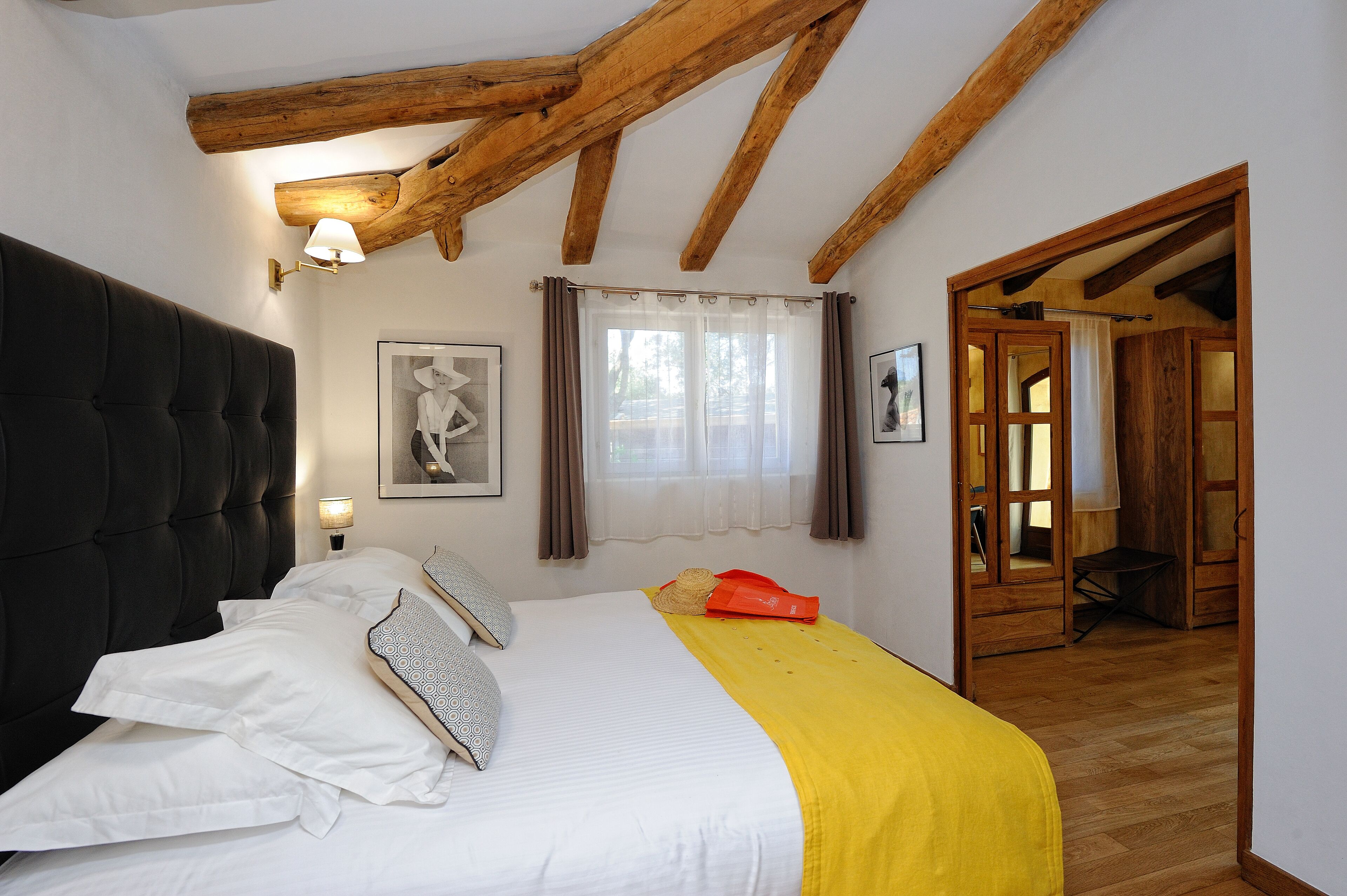 Camera da letto con un letto grande, travi in legno, una finestra con tende e un ingresso che conduce in un'altra stanza.