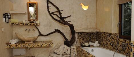 A bathroom with a large bathtub, a sink with a mosaic tile backsplash, a mirror, and a unique tree branch design on the wall.