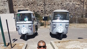 A man sitting on a concrete pathway with two white vehicles in the background. - Casa Marina (Cagliari)