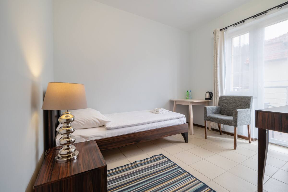 A hotel room with a bed, a lamp, a chair, a small table, and a view of the outside.
