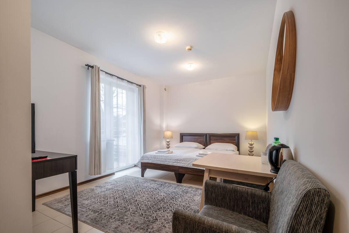 A hotel room with a bed, a desk, a chair, a table, a lamp, and a wall-mounted shelf.
