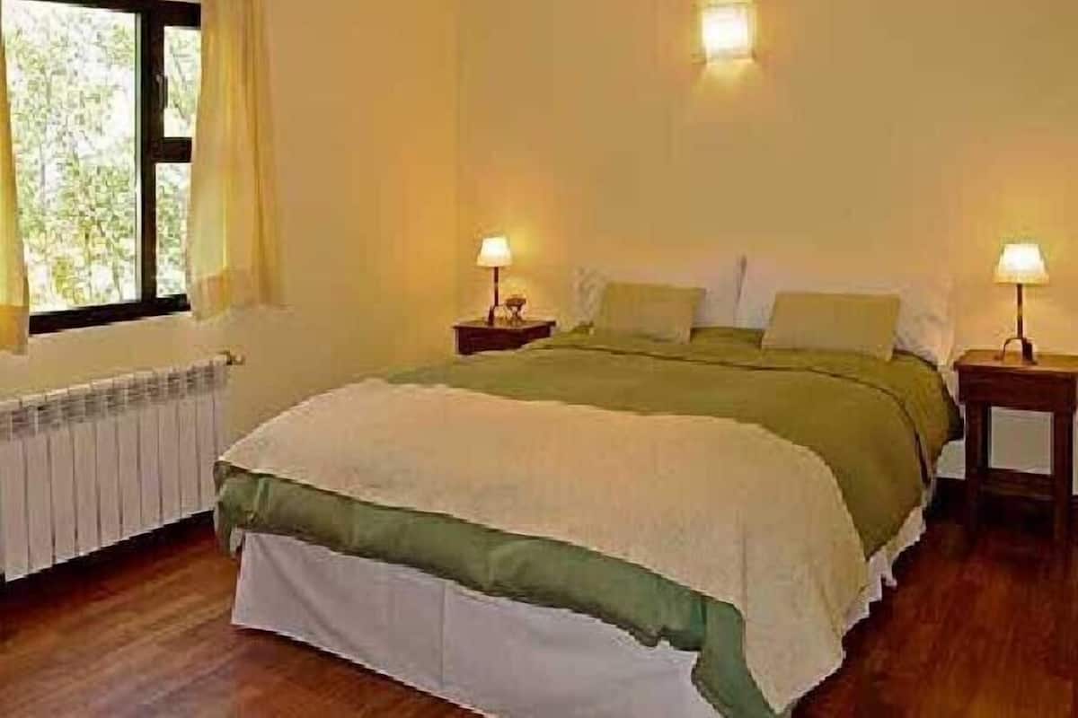 A bedroom with a large bed, two bedside tables with lamps, a radiator, and a window with curtains.