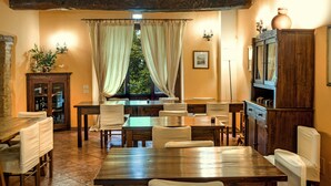 A dining area with wooden tables and chairs, a large window with curtains, and a cabinet with a vase on top. - Agriturismo Sasso Rosso (Assisi)