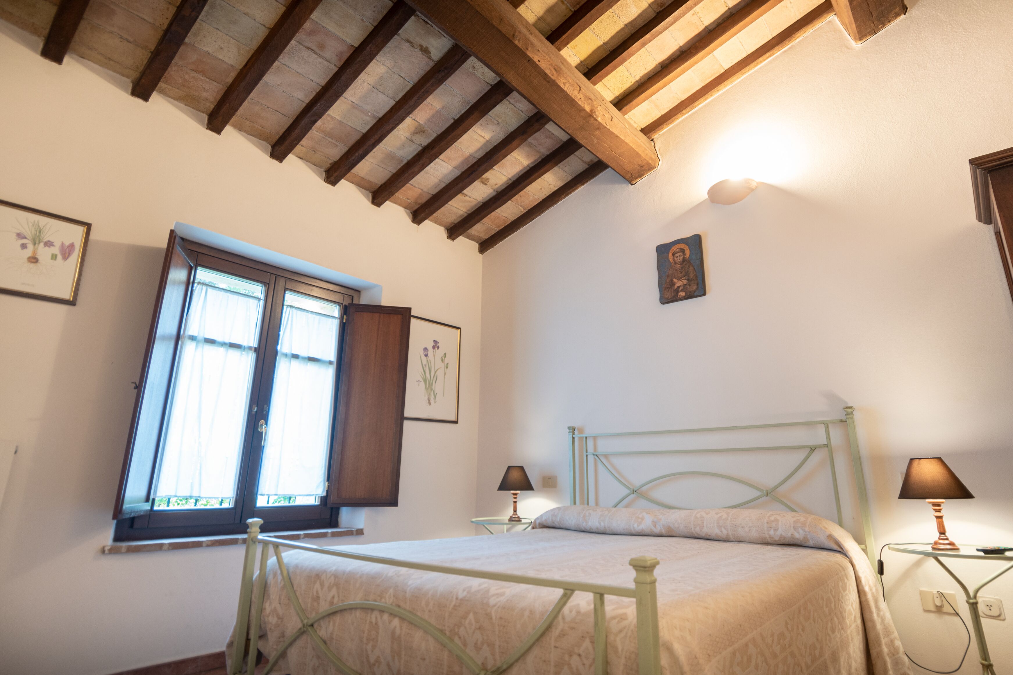 A bedroom with a bed, two bedside lamps, a window with curtains, and wooden beams on the ceiling.