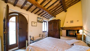 A bedroom with a large bed, wooden ceiling beams, a fireplace, and a door leading outside. - Agriturismo Sasso Rosso (Assisi)
