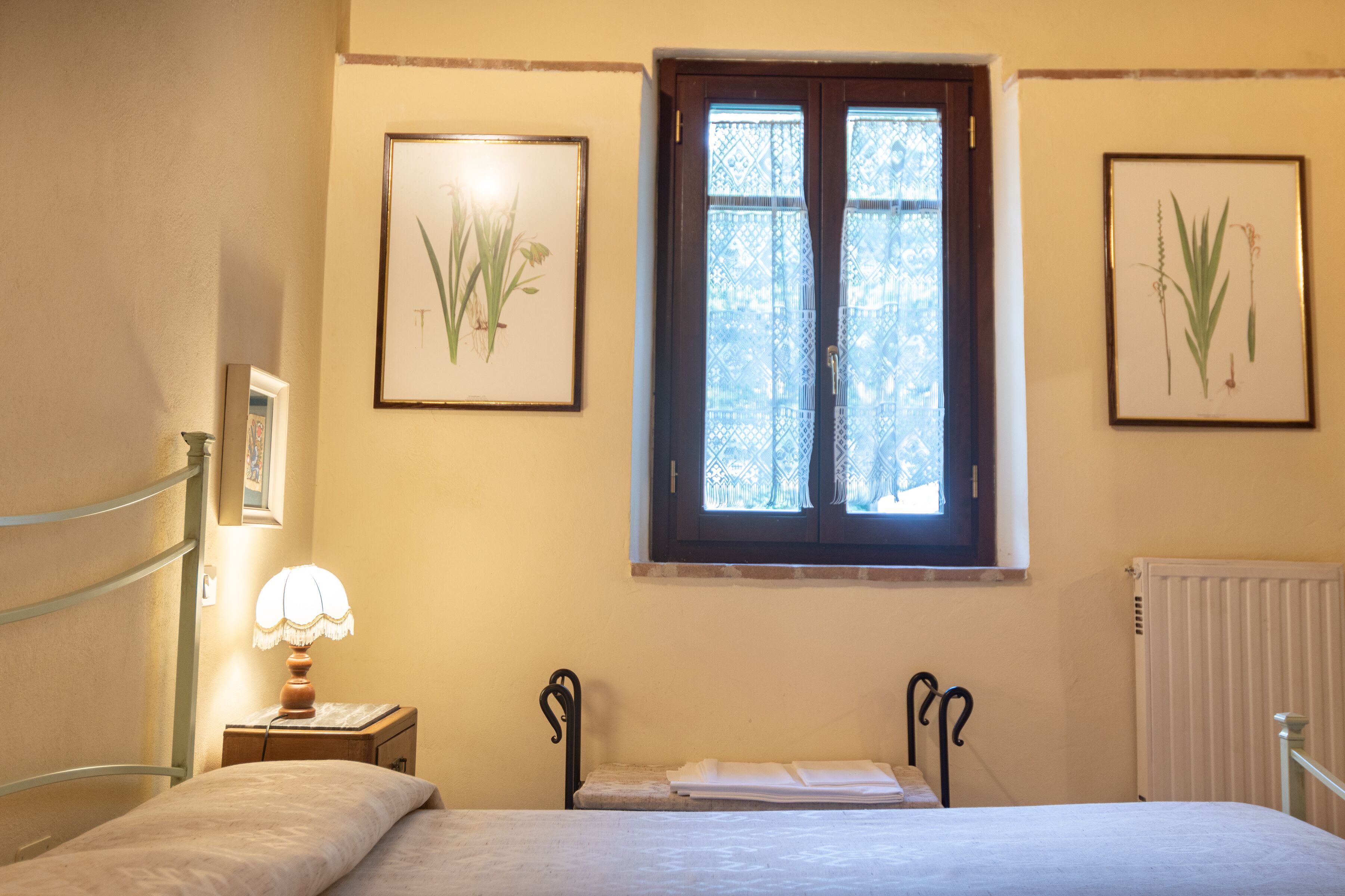 Une chambre à coucher avec un lit, une table de chevet avec une lampe, une fenêtre ornée de rideaux en dentelle et des reproductions encadrées de motifs botaniques accrochées au mur.