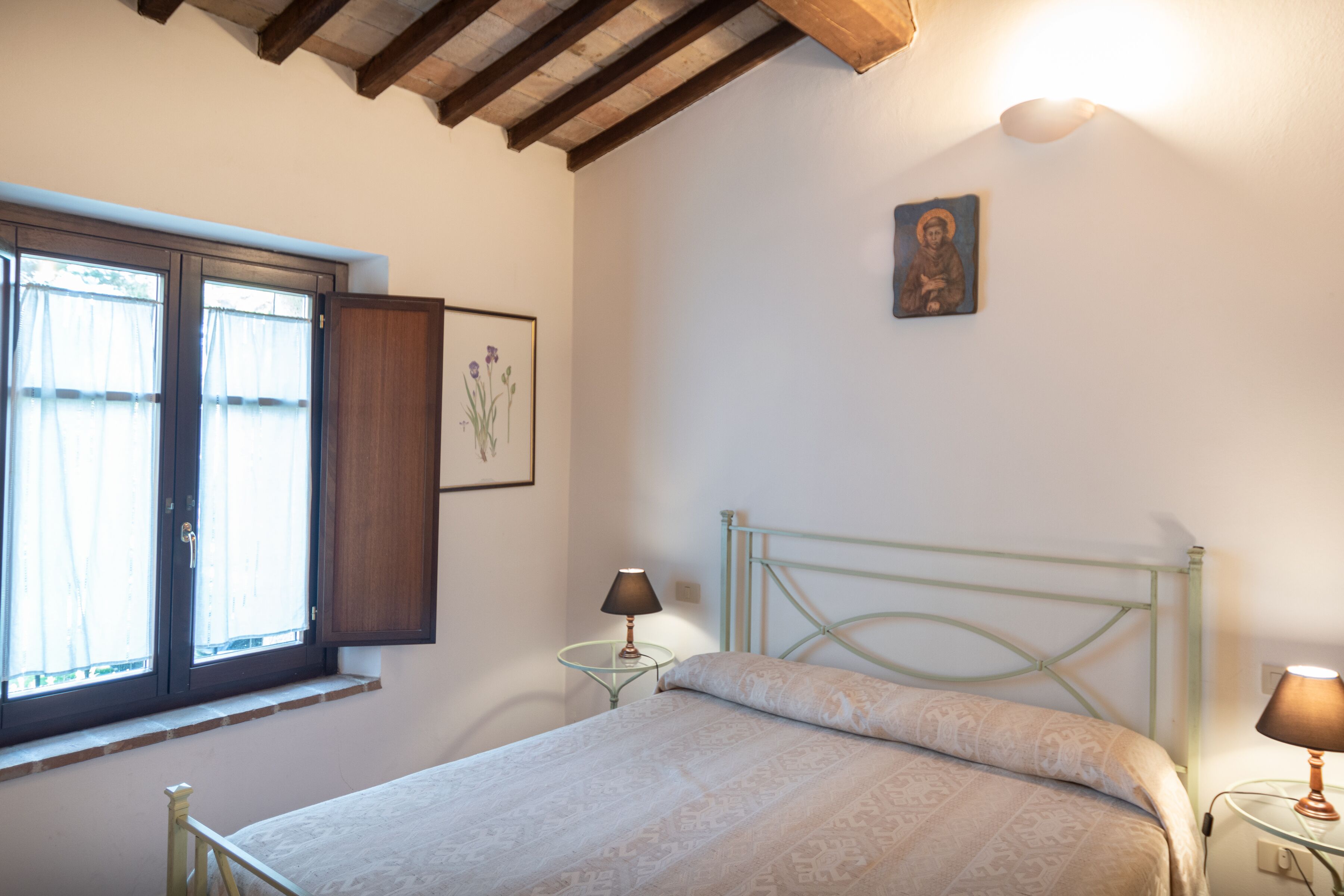 Een slaapkamer met een bed, een raam met gordijnen, een nachtkastje met een lamp en een aan de muur hangend schilderij.