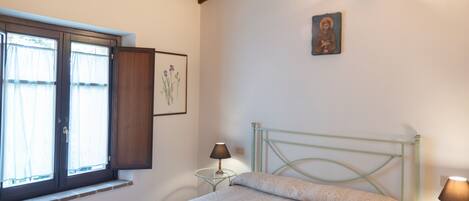 Een slaapkamer met een bed, een raam met gordijnen, een nachtkastje met een lamp en een aan de muur hangend schilderij.
