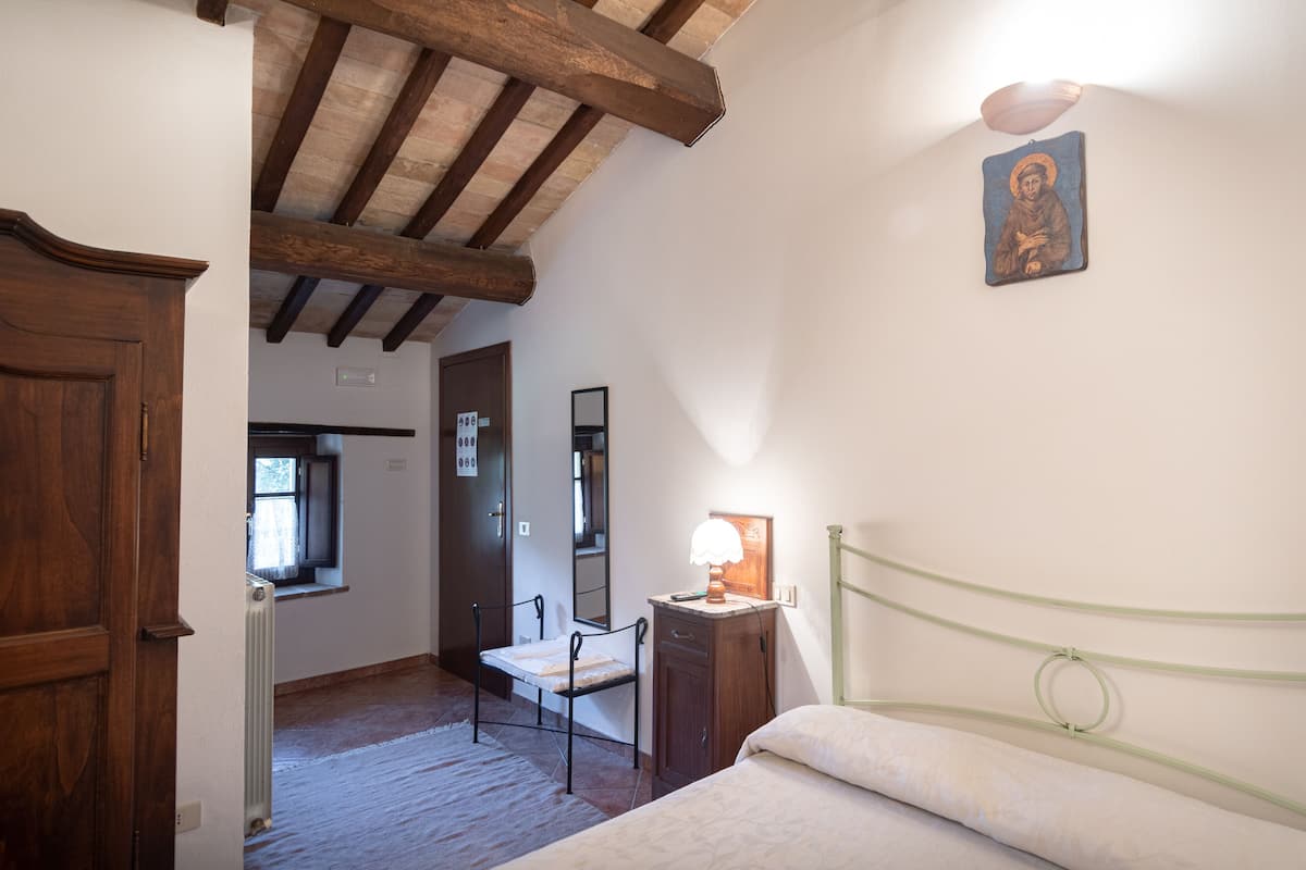 A bedroom with a wooden ceiling, a bed with a green headboard, a nightstand with a lamp, a chair, and a wall-mounted mirror.