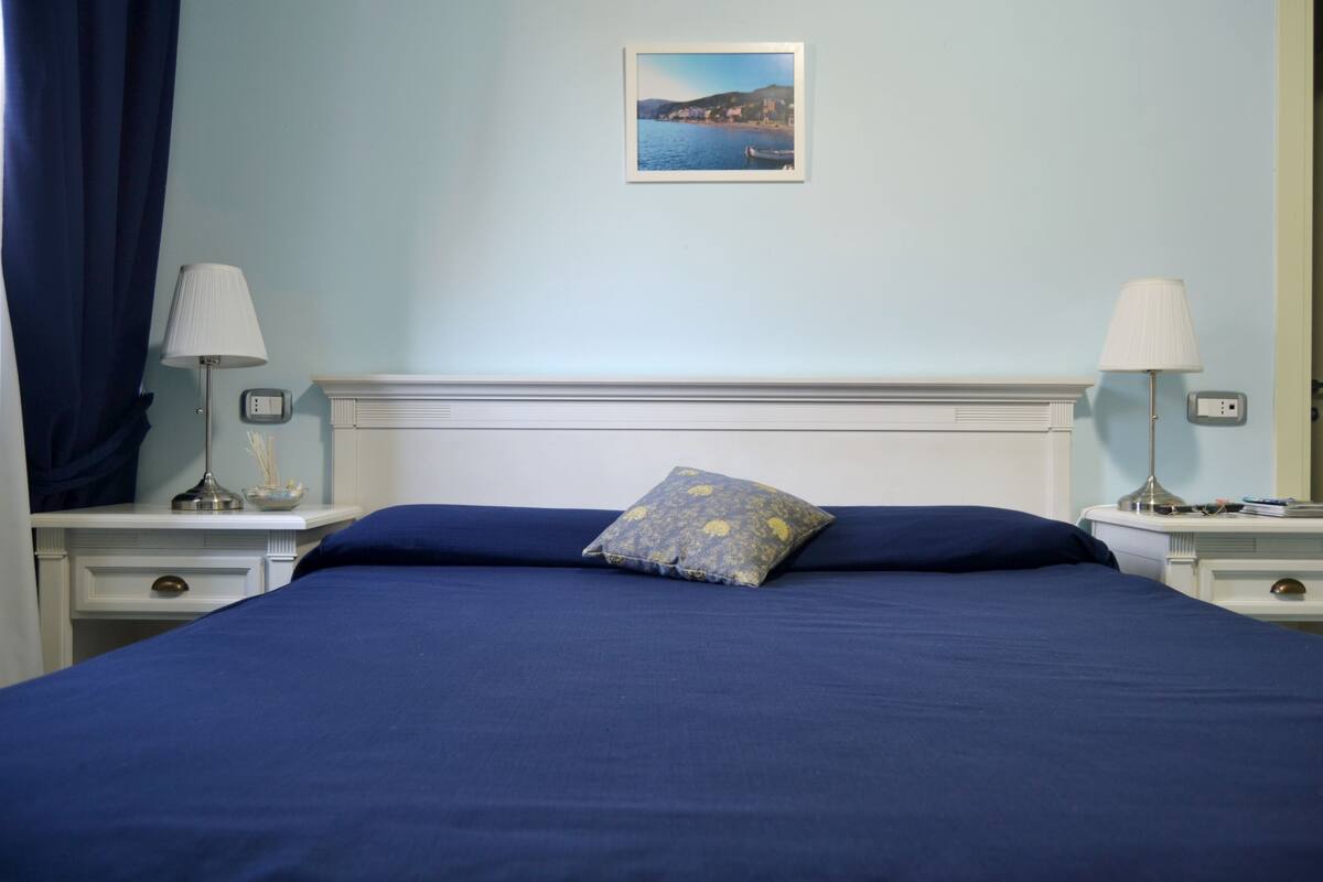 A bed with a blue bedspread and a decorative pillow, flanked by two bedside tables with lamps and a small decorative item on each.