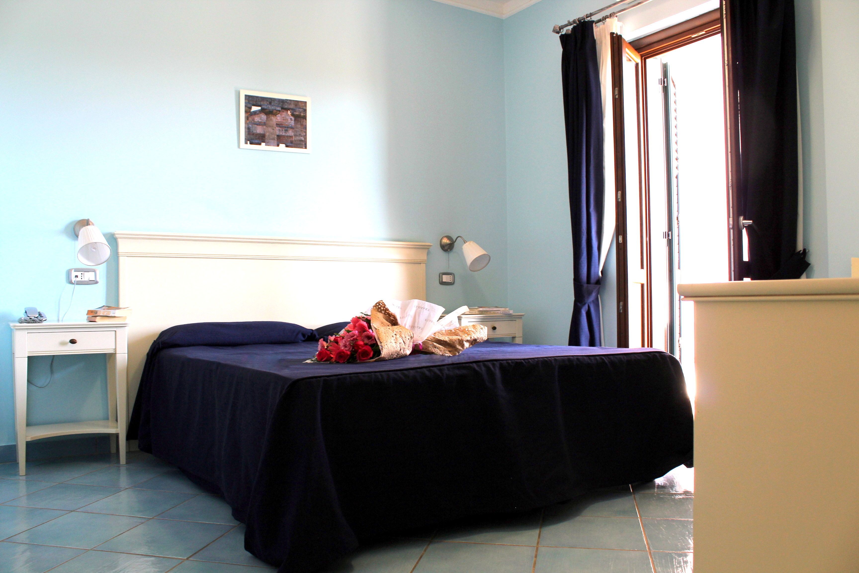 Un dormitorio con cama, mesitas de noche, lámpara, un cuadro en la pared y una ventana con cortinas.