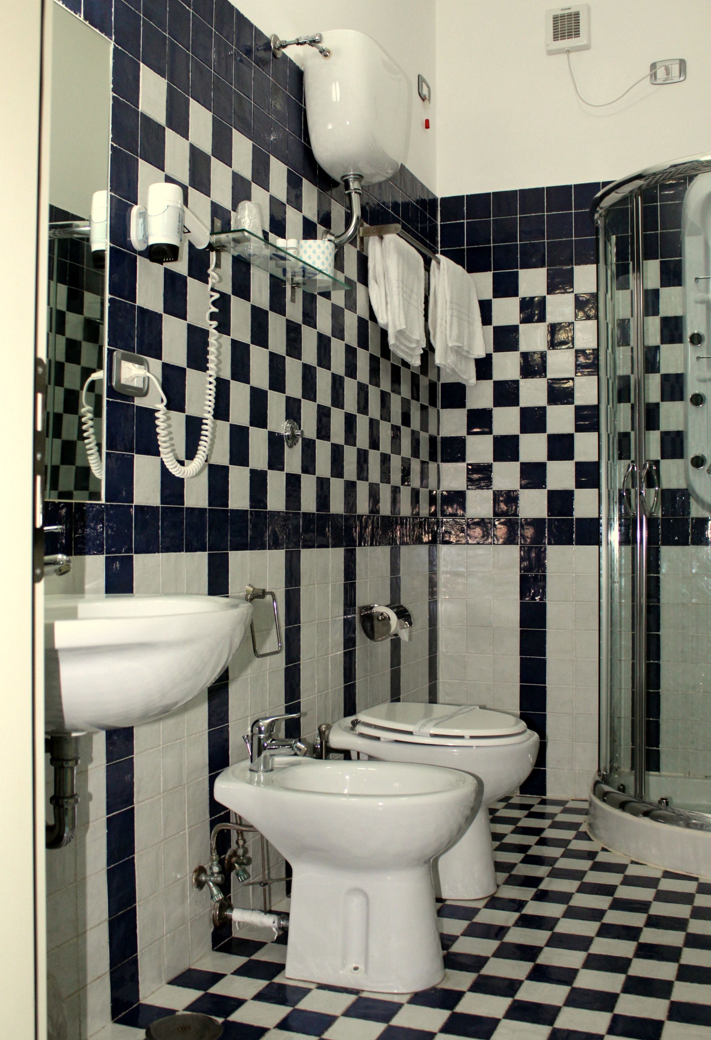 Ein Badezimmer mit Schachbrettboden, einem Waschbecken, zwei Toiletten, einer Dusche und einem Spiegel.