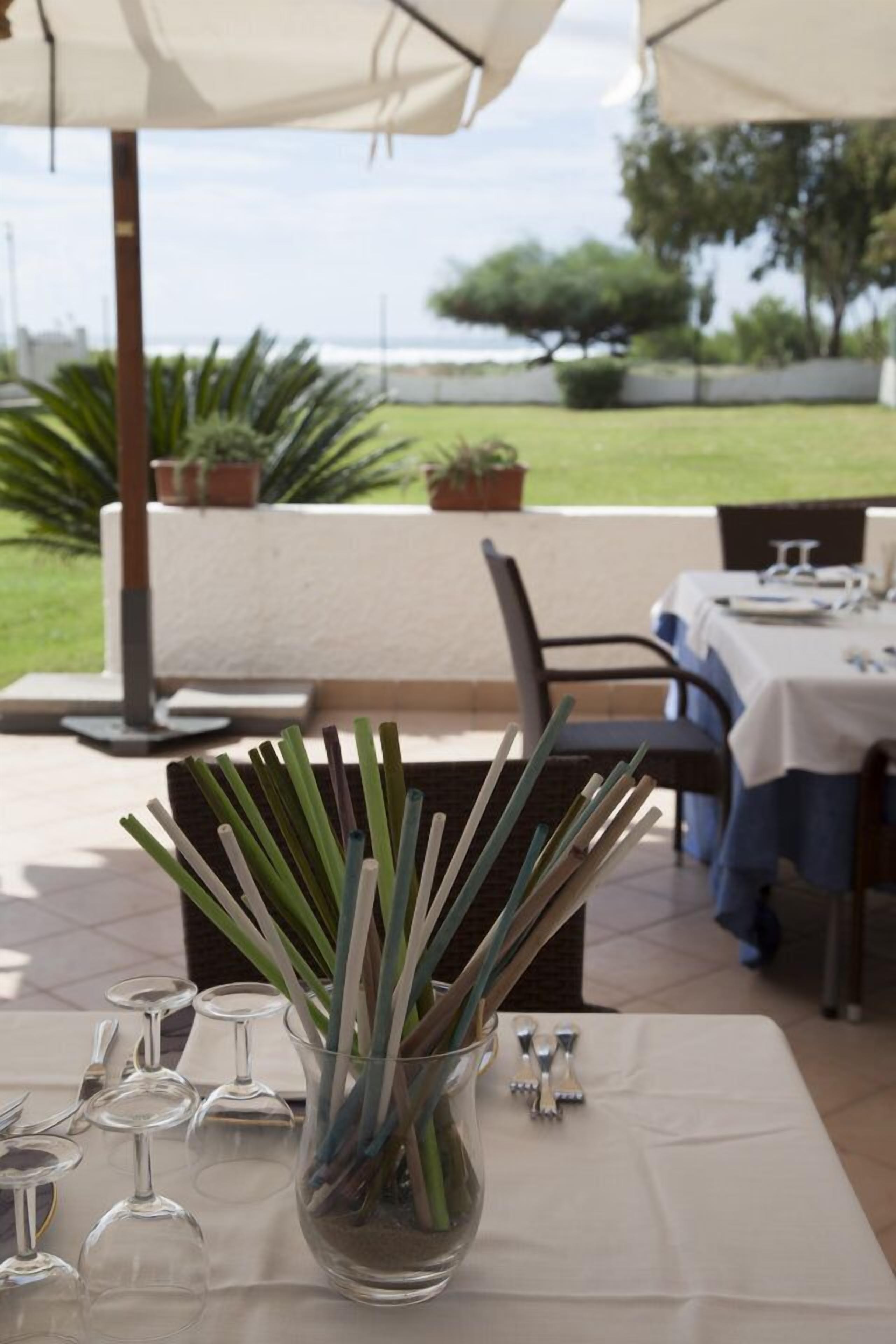 An outdoor dining area with a table set for two, a glass vase with green stems, and a view of the sea and greenery in the background.