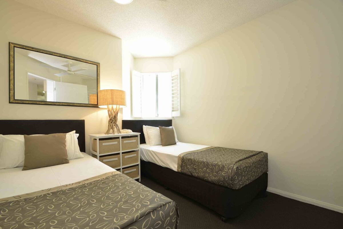 A hotel room with two beds, a nightstand, a lamp, a mirror, and a window with blinds.
