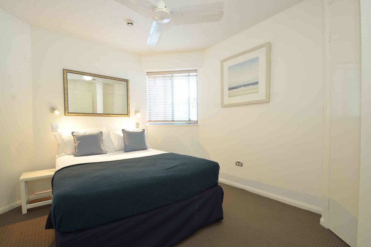 A bedroom with a bed, a nightstand, a mirror, a window, and a framed picture on the wall.