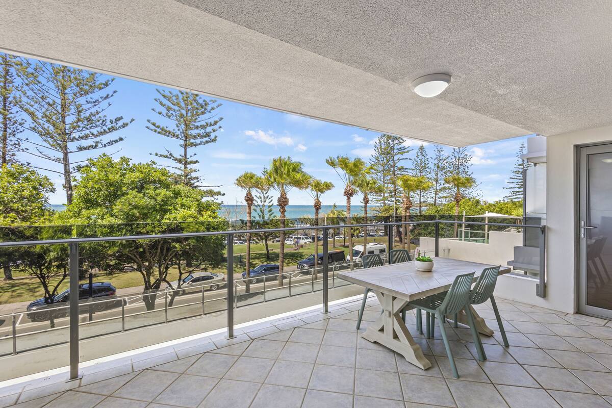 A balcony with a table and chairs, overlooking a street with cars and trees, and a view of the ocean.